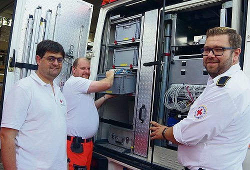 Drei Männer in weißer Kleidung stehen an einem Transporter, dessen Hintertüren geöffnet sind. Man sieht vielen Kisten, die in Schienen hängen. Ein Mann zieht gerade eine Kiste heraus. Alle drei lächeln in die Kamera.