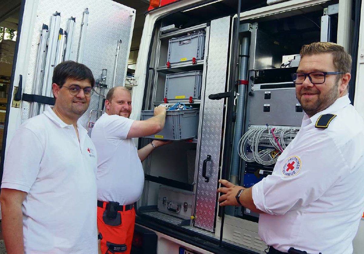 Drei Männer in weißer Kleidung stehen an einem Transporter, dessen Hintertüren geöffnet sind. Man sieht vielen Kisten, die in Schienen hängen. Ein Mann zieht gerade eine Kiste heraus. Alle drei lächeln in die Kamera.