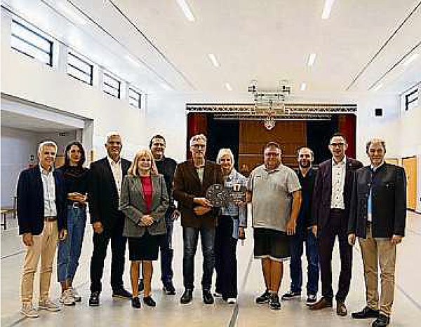 In einer hellen großen Halle steht eine Gruppe von Menschen beieinander. Sie alle lächeln in die Kamera.