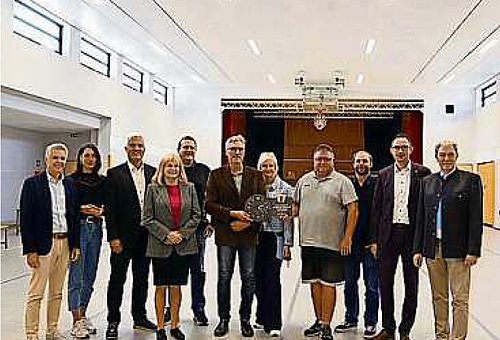 In einer hellen großen Halle steht eine Gruppe von Menschen beieinander. Sie alle lächeln in die Kamera.
