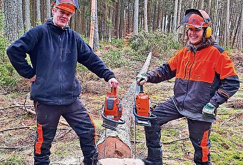 Zwei junge Männer in Sicherheitskleidung mit Motorsägen in der Hand schauen lächeln in die Kamera.