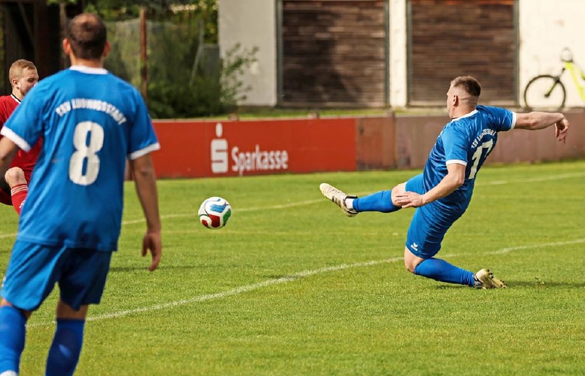 Zwei Fußballspieler in blauen Trikots auf einem grünen Fußballplatz. Einer der Spieler tritt gerade den Ball. 