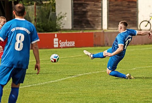 Zwei Fußballspieler in blauen Trikots auf einem grünen Fußballplatz. Einer der Spieler tritt gerade den Ball. 