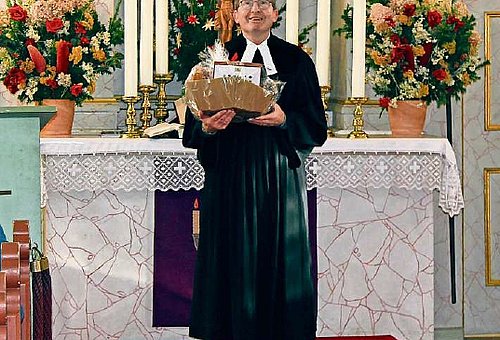 Ein älterer Mann im Talar steht mit einem Geschenkkorb vor dem Altar in einer Kirche.