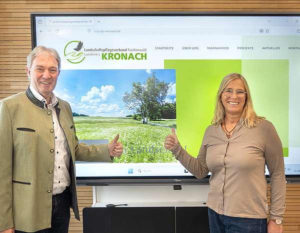 Eine Frau und ein Mann stehen vor einer Leinwand. Darauf ist die Seite einer Website zu sehen. Beide lächeln in die Kamera.