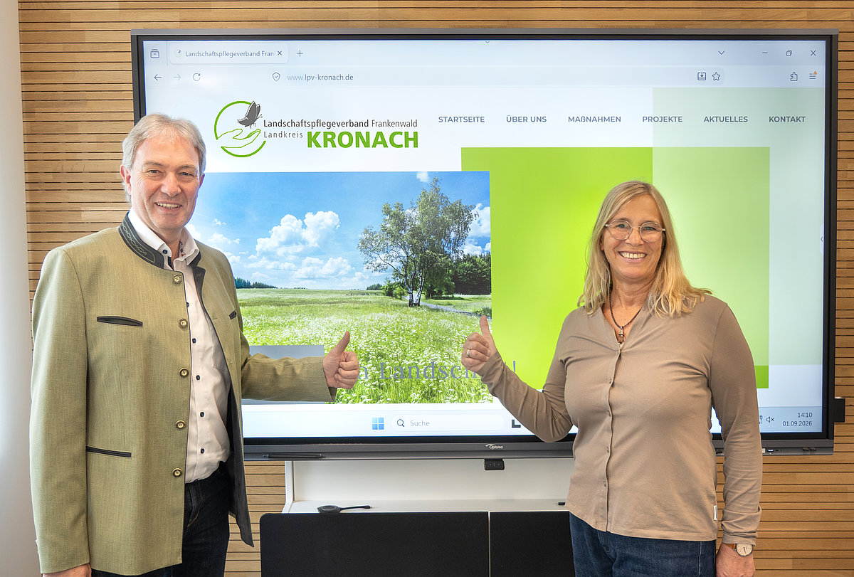 Eine Frau und ein Mann stehen vor einer Leinwand. Darauf ist die Seite einer Website zu sehen. Beide lächeln in die Kamera.