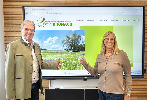 Eine Frau und ein Mann stehen vor einer Leinwand. Darauf ist die Seite einer Website zu sehen. Beide lächeln in die Kamera. 