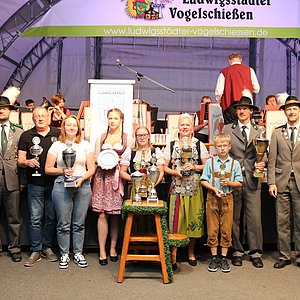 Vor einer Bühne in einem Zelt steht ein Gruppe vo Menschen verschiednen Alters. Einige tragen Dirndl und Schützenuniformen und einige tragen Freizeitkleidung. Ein paar halten je einen Pokal in ihren Händen.