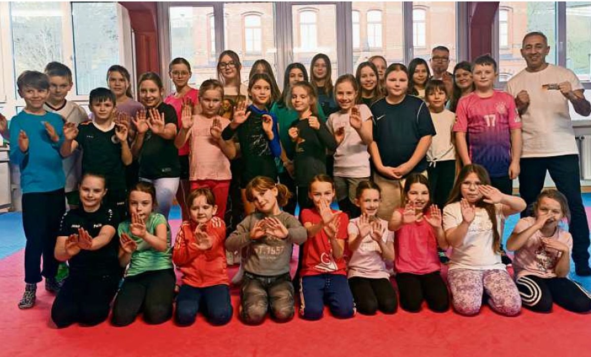 Eine große Gruppe Kinder stehen und hocken in mehreren Reihen hintereinander. Alle tragen Sport- oder Freizeitkleidung und machen mit ihren Hände eine Abwehrpose. Daneben steht der Bundestrainer Harun Elkol.