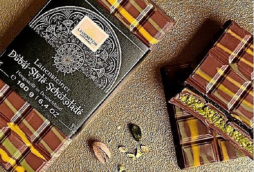 Produktbild der beschriebenen Schokolade. Eine komplette Tafel mit schwarzer Banderole liegt neben zwei angebrochenen Teilstücken einer Tafel.