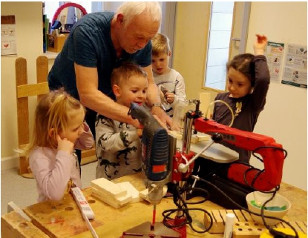 In einer kleinen hellen Werkstatt stehen vier Kinder um eine Werkbank herum. Sie schauen interessiert. Ein älterer Mann mit grauen Haaren hilft einem Kind bei der Bedienung einer Standbohrmaschine.