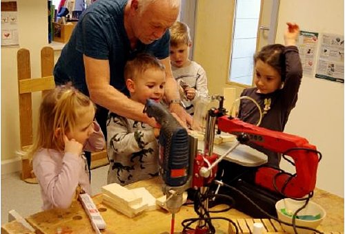 In einer kleinen hellen Werkstatt stehen vier Kinder um eine Werkbank herum. Sie schauen interessiert. Ein älterer Mann mit grauen Haaren hilft einem Kind bei der Bedienung einer Standbohrmaschine.