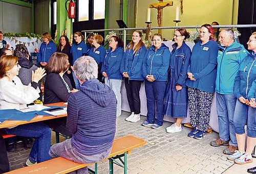 In einem Raum sitzen Menschen an Biertischgarnituren. Ganz vorn stehen Frauen in blauen Shirts in einer Reihe nebeneinander. Die Menschen klatschen für die Frauen.