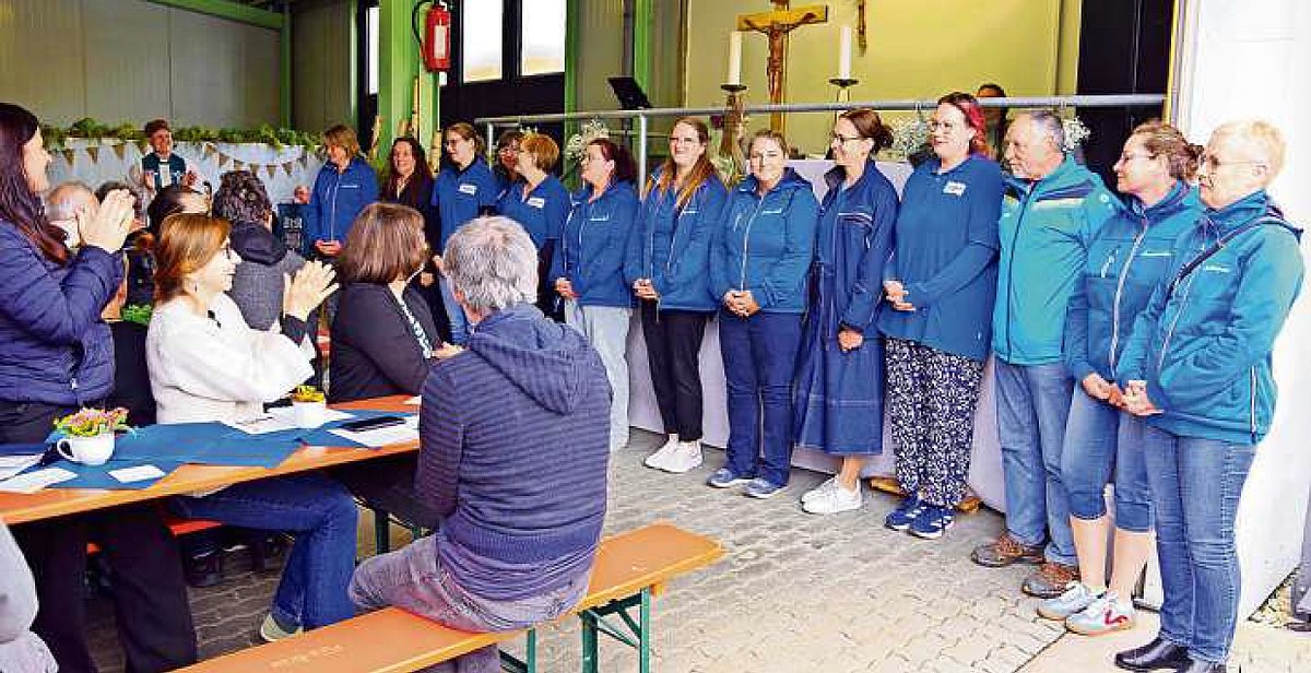 In einem Raum sitzen Menschen an Biertischgarnituren. Ganz vorn stehen Frauen in blauen Shirts in einer Reihe nebeneinander. Die Menschen klatschen für die Frauen.