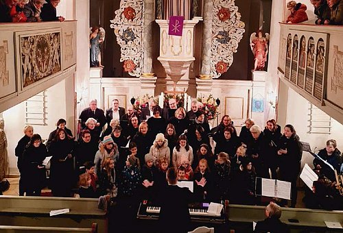 Im Inneren einer Kirche vor dem Altar stehen vielen Frauen, Männer und Kinder in schwarzer Kleidung und schauen zu ihrem Dirigenten.