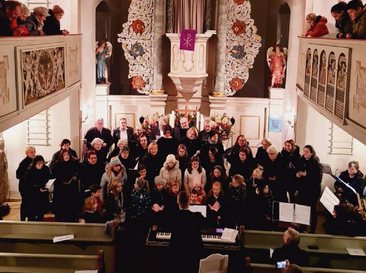 Im Inneren einer Kirche vor dem Altar stehen vielen Frauen, Männer und Kinder in schwarzer Kleidung und schauen zu ihrem Dirigenten.