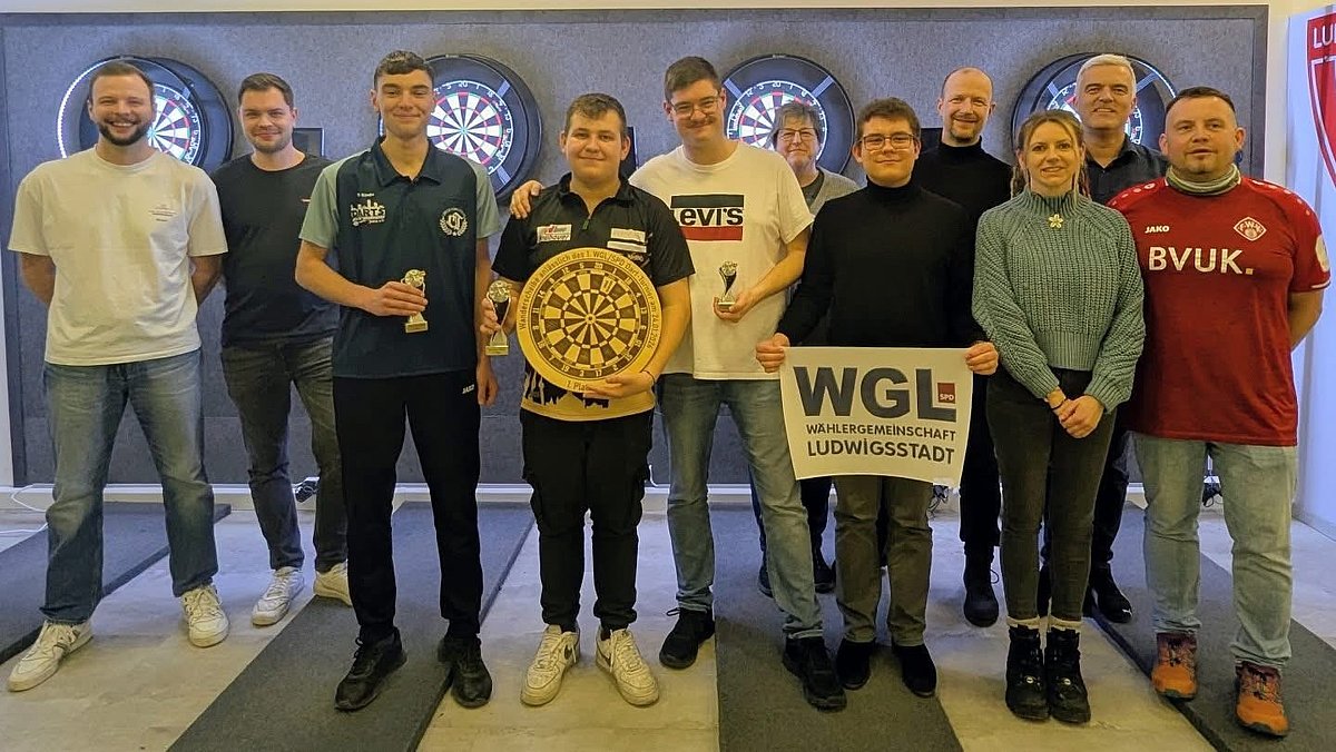 Vor einer Wand mit vier Dartscheiben stehen elf Personen in Alltagskleidung. In der Mitte steht ein junger Mann mit einer Dartscheibe und einem Pokal in seinen Händen. Rechts und links ebenfalls zwei Männer mit Pokalen. Alle lächeln in die Kamera.