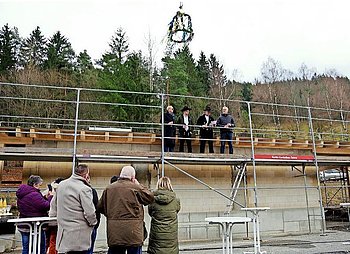 Vier Personen stehen auf einem Gerüst, davon hält ein Zimmermann in seiner Kluft den Richtspruch. Über ihnen schwebt die Richtkrone. Ihnen zugewandt eine Gruppe, die bei diesem Festakt dabei waren.
