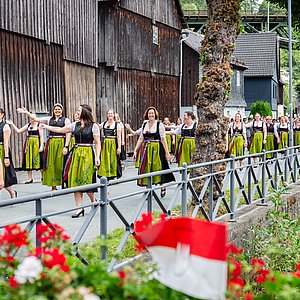 Entlang einer geschmückten Straße laufen zahlreiche Frauen in Dirndln. Sie tragen schwarze Kleider mit weißen Blusen und Schürzen, die grün und pink sind. Alle lachen und winken.