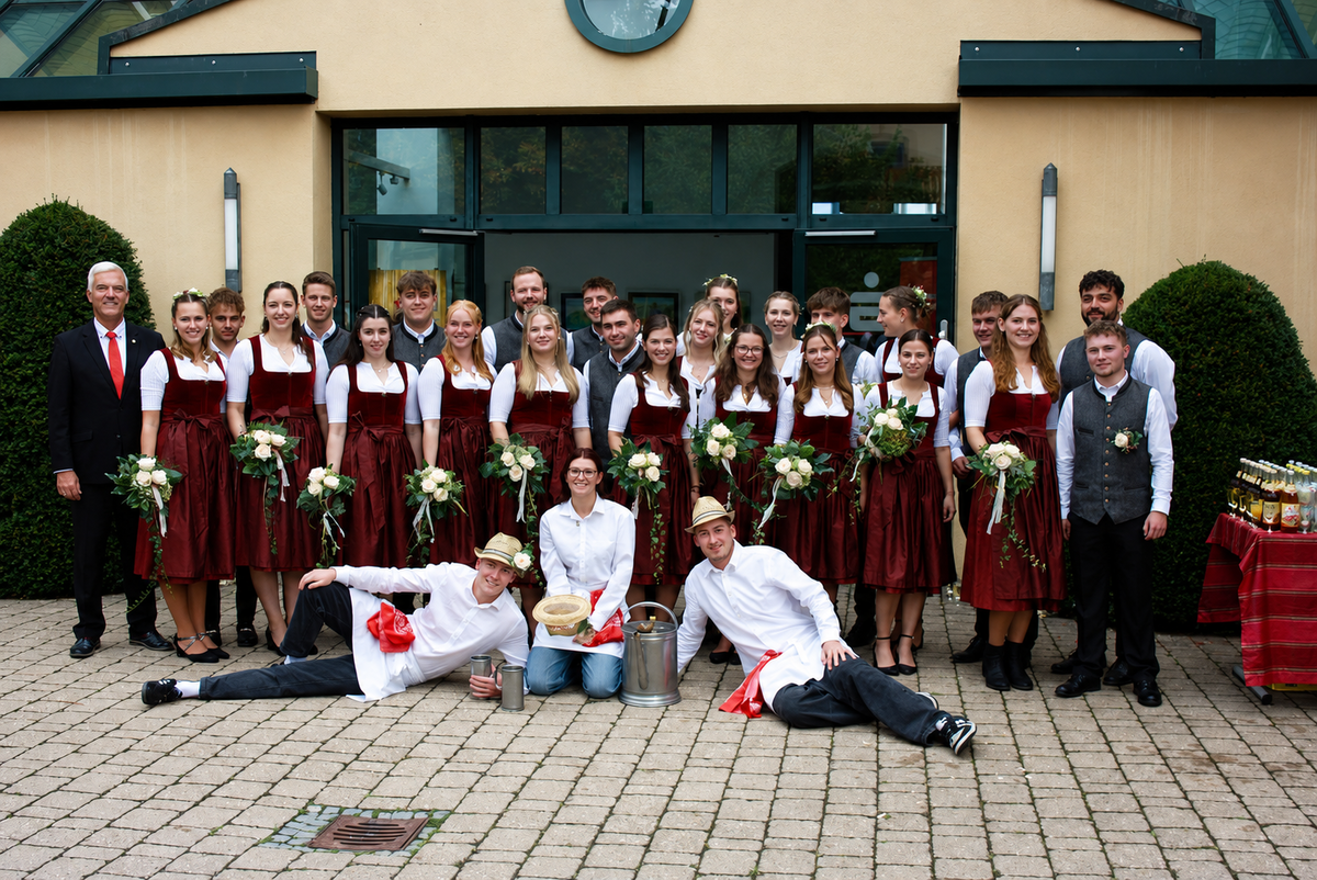 Vor einem Gebäude ist eine große Gruppe von jungen Menschen. Alle Mädchen tragen dunkelrote Dirndl und halten einen kleinen Blumenstrauß in der Hand. Die Burschen tragen dunkle Hosen, weiße Hemden und graue Westen. Am linken Rand steht der Erste Bürgermeister und vor der Gruppe liegen die sogenannten Kellner. Das sind junge Männer mit dunklen Hosen, weißen Hemden und Schürzen sowie Hüten. Alle lächeln in die Kamera.