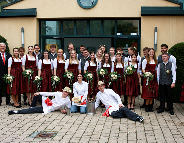 Vor einem Gebäude ist eine große Gruppe von jungen Menschen. Alle Mädchen tragen dunkelrote Dirndl und halten einen kleinen Blumenstrauß in der Hand. Die Burschen tragen dunkle Hosen, weiße Hemden und graue Westen. Am linken Rand steht der Erste Bürgermeister und vor der Gruppe liegen die sogenannten Kellner. Das sind junge Männer mit dunklen Hosen, weißen Hemden und Schürzen sowie Hüten. Alle lächeln in die Kamera.