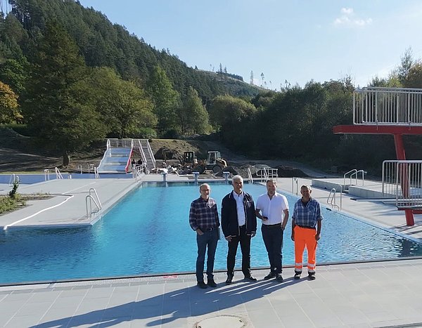 Vor einem gefüllten Freibad-Schwimmerbecken stehen vier Männer. Sie stehen mit dem Rücken zum Becken und schauen in die Kamera.