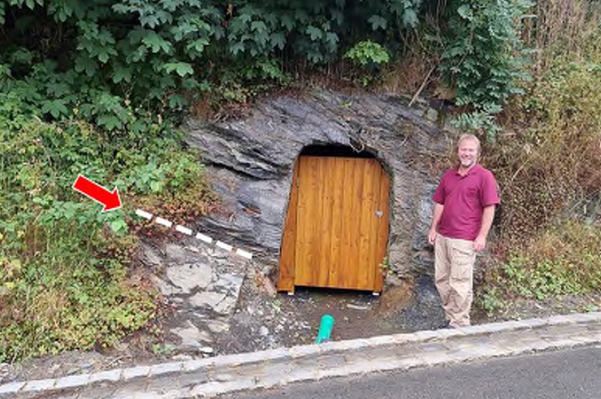 Ein Mann steht an einer Felswand neben dem Eingang zu einem Felsenkeller, der mit einer Holzstür verschlossen ist.