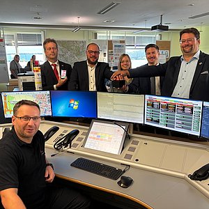 Ein Mann sitzt an einem Computerarbeitsplatz mit vielen Monitoren. An dem Schreibtisch hinter den Bildschirmen stehen vier Männer und in deren Mitte eine Frau. Alle lächeln in die Kamera.