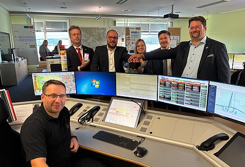 Ein Mann sitzt an einem Computerarbeitsplatz mit vielen Monitoren. An dem Schreibtisch hinter den Bildschirmen stehen vier Männer und in deren Mitte eine Frau. Alle lächeln in die Kamera.