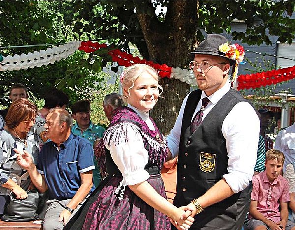 Im Fokus des Bildes ein Tanzpaar in Tanzhaltung. Sie stehen zueinander und halten sich an den Händen. Der Mann trägt ein weißes Hemd dazu eine dunkle Weste, eine dunkle Hose und einen dunklen Hut. An dem Hut steckt eine buntes Blumensträußchen. Seine Tanzpartnerin trägt eine weiße Bluse und ein dunkles Dirndl mit einem lila Tuch und einer lila Schürze. Beide lächeln - im Hintergrund sind Zuschauer zu sehen. 