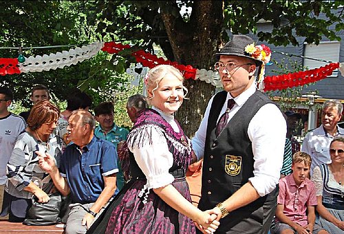 Im Fokus des Bildes ein Tanzpaar in Tanzhaltung. Sie stehen zueinander und halten sich an den Händen. Der Mann trägt ein weißes Hemd dazu eine dunkle Weste, eine dunkle Hose und einen dunklen Hut. An dem Hut steckt eine buntes Blumensträußchen. Seine Tanzpartnerin trägt eine weiße Bluse und ein dunkles Dirndl mit einem lila Tuch und einer lila Schürze. Beide lächeln - im Hintergrund sind Zuschauer zu sehen. 