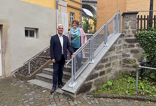 Ein Mann im Anzug und eine Frau stehen draußen auf einer Treppe mit silbergrauem Geländer. Beide schauen in die Kamera.