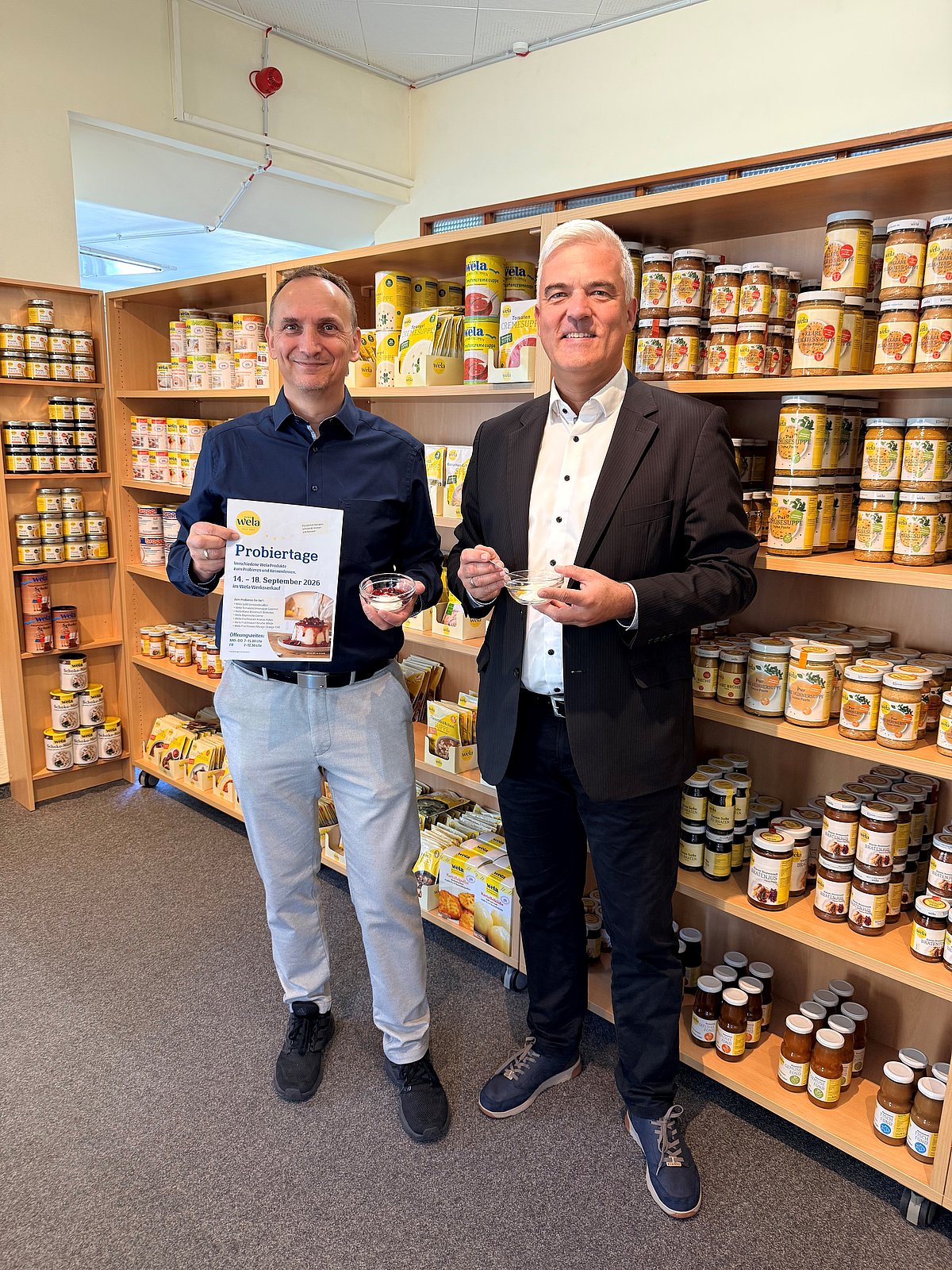 Zwei Männer befinden sich im Verkaufsraum der Wela-Werke. Sie halten ein Schüsselchen mit Süßspeise in der Hand. In den Regalen hinter ihnen sind verschiedene Produkte zu sehen.