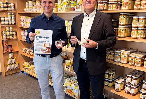 Zwei Männer befinden sich im Verkaufsraum der Wela-Werke. Sie halten ein Schüsselchen mit Süßspeise in der Hand. In den Regalen hinter ihnen sind verschiedene Produkte zu sehen.