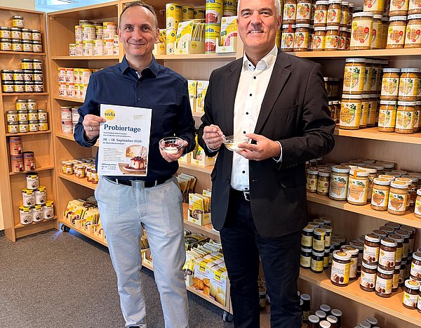 Zwei Männer befinden sich im Verkaufsraum der Wela-Werke. Sie halten ein Schüsselchen mit Süßspeise in der Hand. In den Regalen hinter ihnen sind verschiedene Produkte zu sehen.