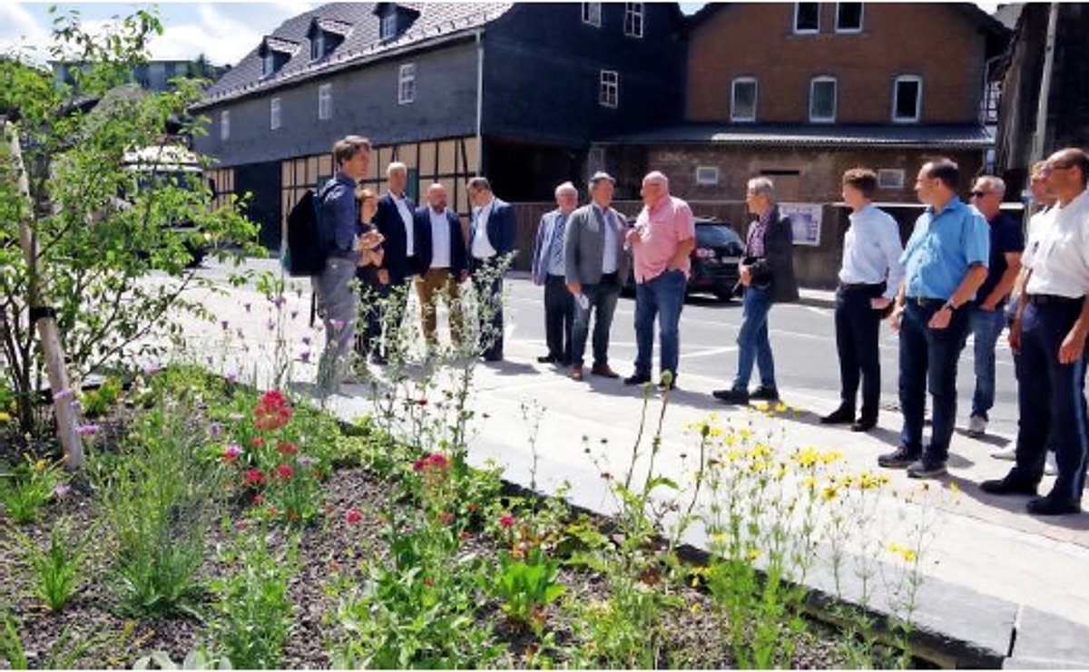 Eine Gruppe von Menschen steht an einer Straße zusammen. Vor ihnen befinden sich Blumenbeete. Hinter ihnen die Straße und Häuser.