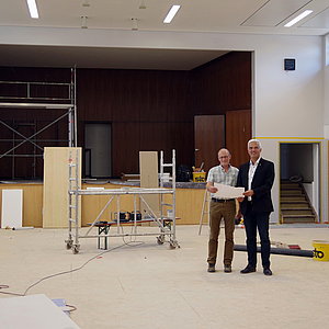 Zwei Männer mit einem Plan in den Händen stehen in einem großen Saal mit Bühne. Der Raum ist eine Baustelle. Es stehen und liegen Bauutensilien überall herum.