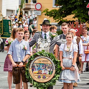 Das Bild zeigt in der Mitte den Schützenkönig, der hinter seiner Schützenscheibe steht. Diese wird von seinen beiden Kindern Jannes und Mara getragen. Jannes trägt eine Lederhose, Mala ein Dirndl.