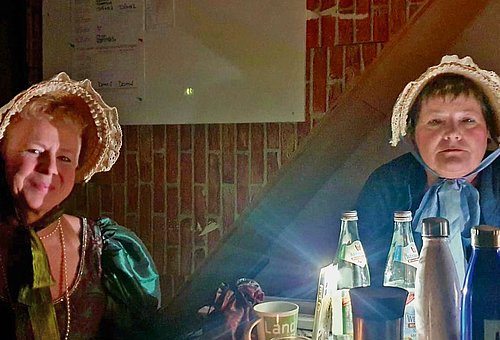 Zwei Frauen sitzen mit ihren Theaterkostümen bei Taschenlampenlicht am Tisch und schauen in die Kamera. 