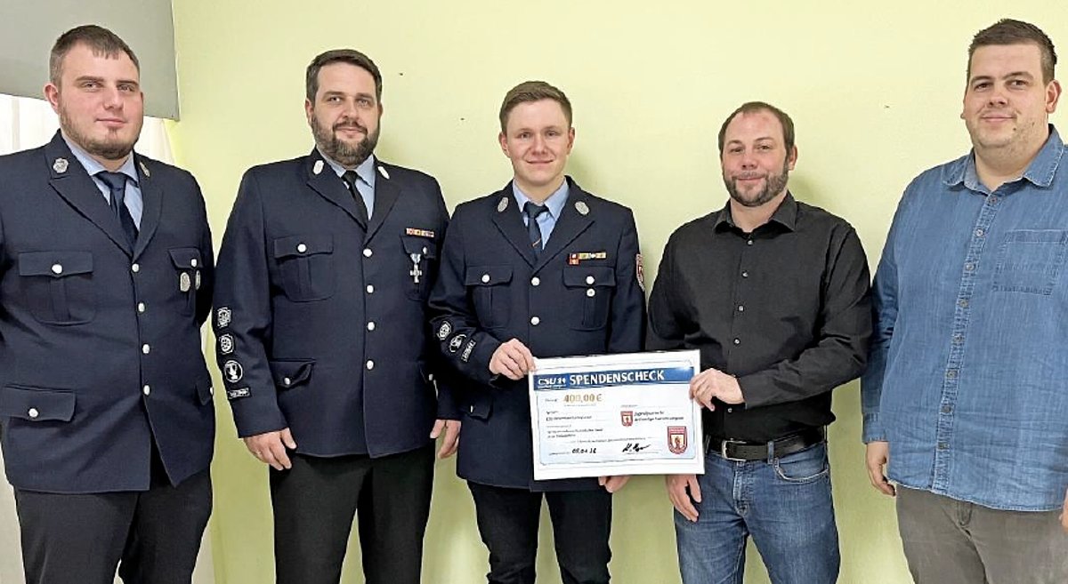 Drei Männer in Feuerwehruniform und zwei Männer in Freizeitkleidung stehen in einer Gruppe. Alle lächeln in die Kamera. Ein Feuwehrmann und einer der beiden anderen Männer halten gemeinsam einen großen Spendenscheck in der Hand.