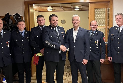 7 Männer stehen in einer Reihe nebeneinander und schauen in die Kamera. Sieben von Ihnen tragen ihre Feuerwehruniform, das sind dunkelblaue Jacken mit Orden und Hosen. Der achte Mann, Bürgermeister Timo Ehrhardt, trägt einen dunklen Anzug.