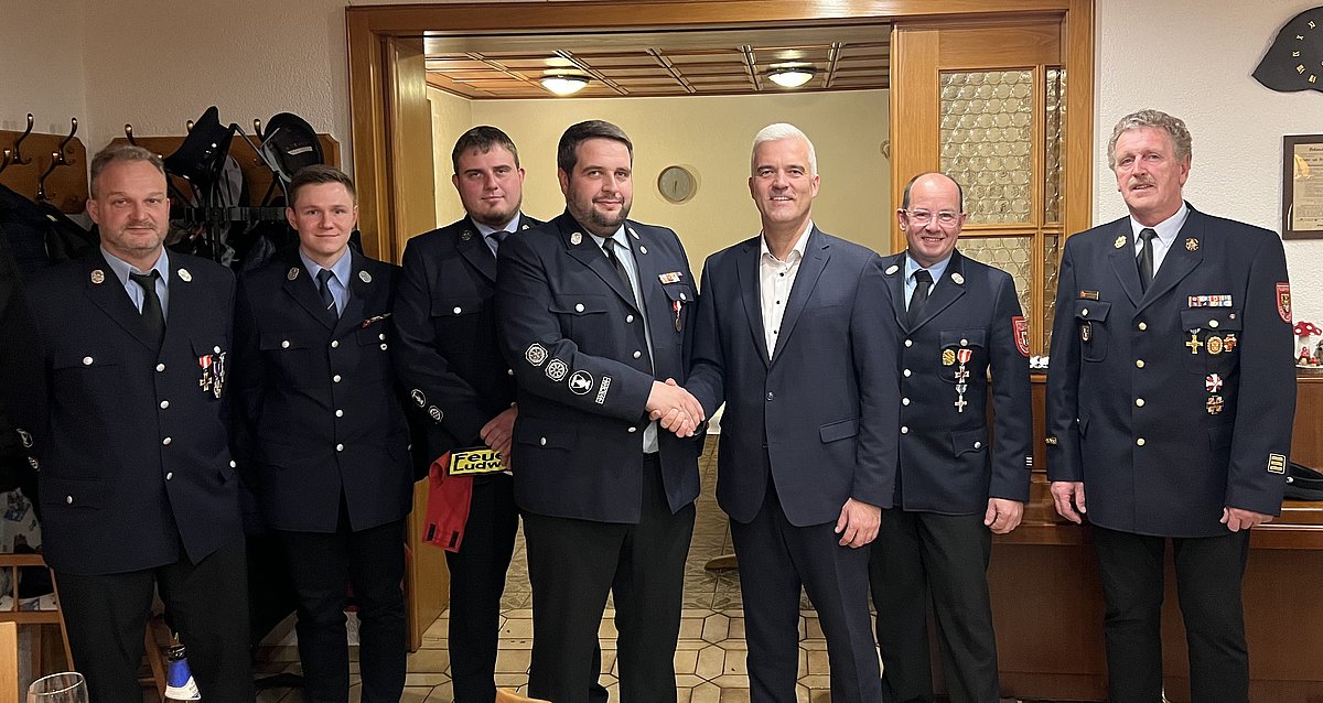 7 Männer stehen in einer Reihe nebeneinander und schauen in die Kamera. Sieben von Ihnen tragen ihre Feuerwehruniform, das sind dunkelblaue Jacken mit Orden und Hosen. Der achte Mann, Bürgermeister Timo Ehrhardt, trägt einen dunklen Anzug.