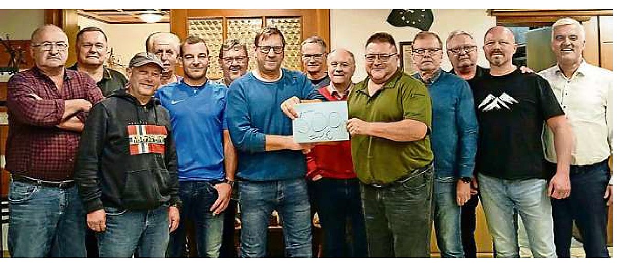 In einem Raum steht eine Gruppe von Menschen zusammen. Alle lächeln in die Kamera. Zwei Männer in der vorderen Reihe halten gemeinsam einen Zettel auf dem 500 € steht.