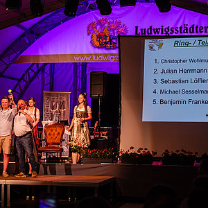 Im Festzelt. Es hängt eine Leinwand, auf der die Ergebnisse des Schießens stehen. Neben der Leinwand auf der Bühne stehen mehrere Personen. Einer davon hat die Arme in die Luft gestreckt.