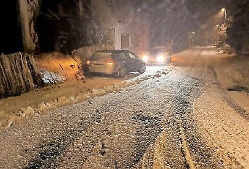 Nachts auf einer schneebedeckten Straße. Ein Auto steht am Straßenrand an einem zerstörten Gartenzaun. Davor sind die Scheinwerfer eines Polizeiautos zu sehen.