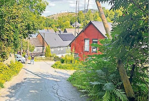 Eine unbefestigte Straße mit Bäumen und Häusern. Im Bild ist eine durchhängende Stromleitung zu sehen. 