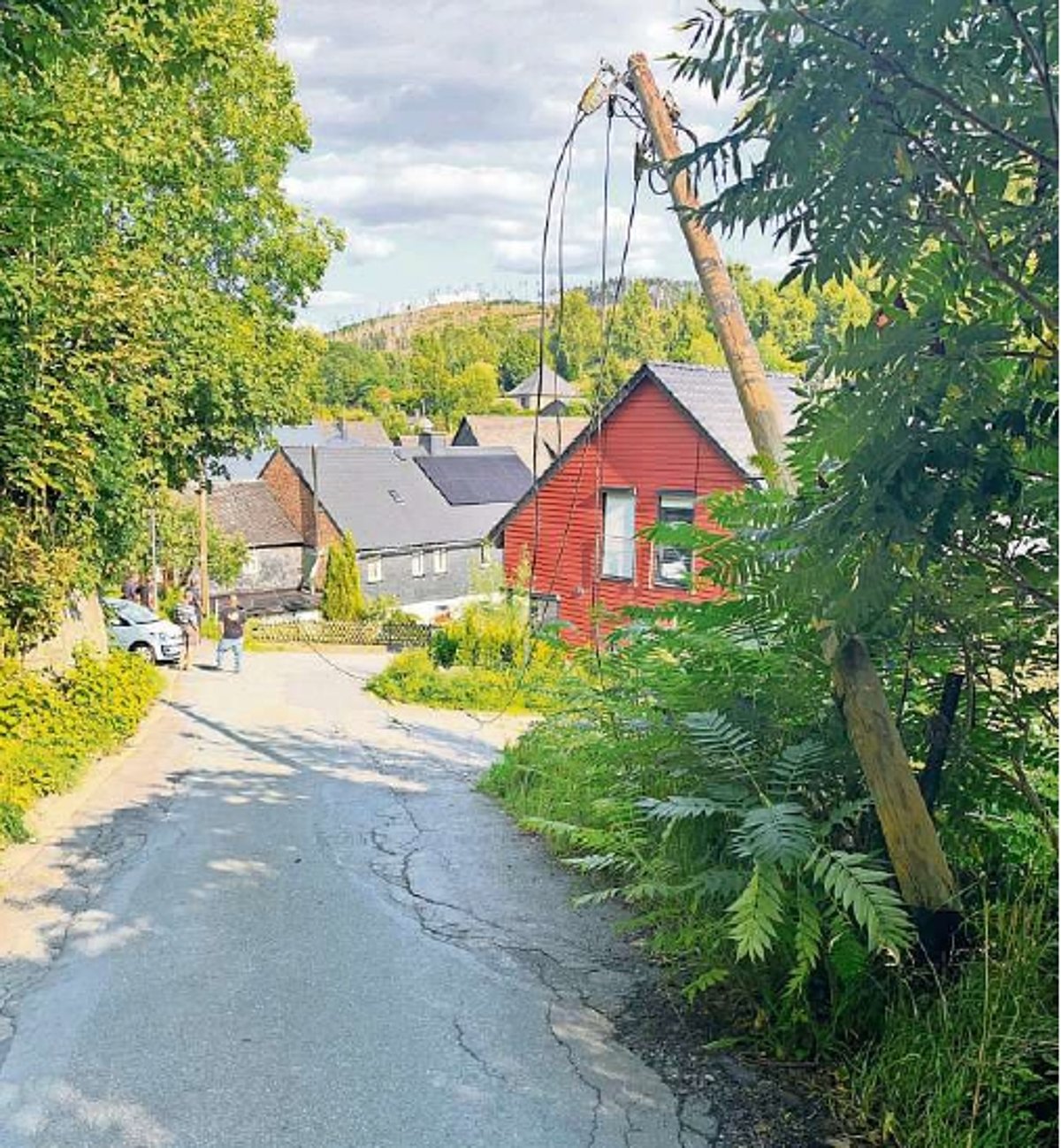 Eine unbefestigte Straße mit Bäumen und Häusern. Im Bild ist eine durchhängende Stromleitung zu sehen.