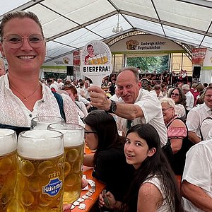 Im Vordergrund eine Bedienung im Dirndl mit vier Maßkrügen Bier. Im Hintergrund hält ein Gast ein Schild mit dem Wort "Bierfraa" in die Luft.