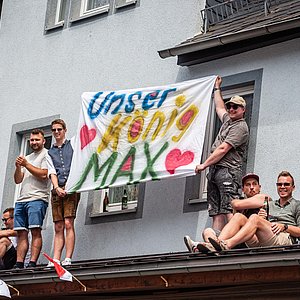 Einige junge Männer sitzen und stehen auf einem Dach und halten ein Transparent auf dem "Unser König Max" steht.