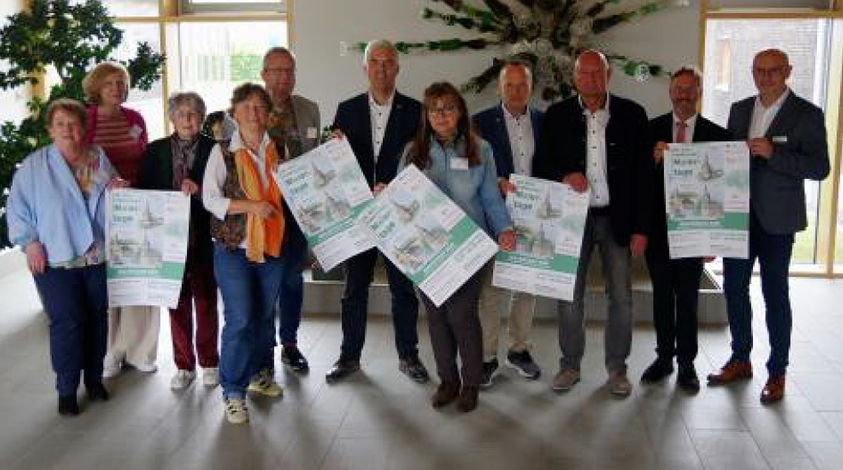 Eine Gruppe von Menschen mit Frauen und Männern in Freizeitkleidung. Sie stehen in einer Gruppe in einem Gebäude. Die Menschen in der ersten Reihe halten Plakate in die Kamera. Sie alle lächeln und schauen in die Kamera.
